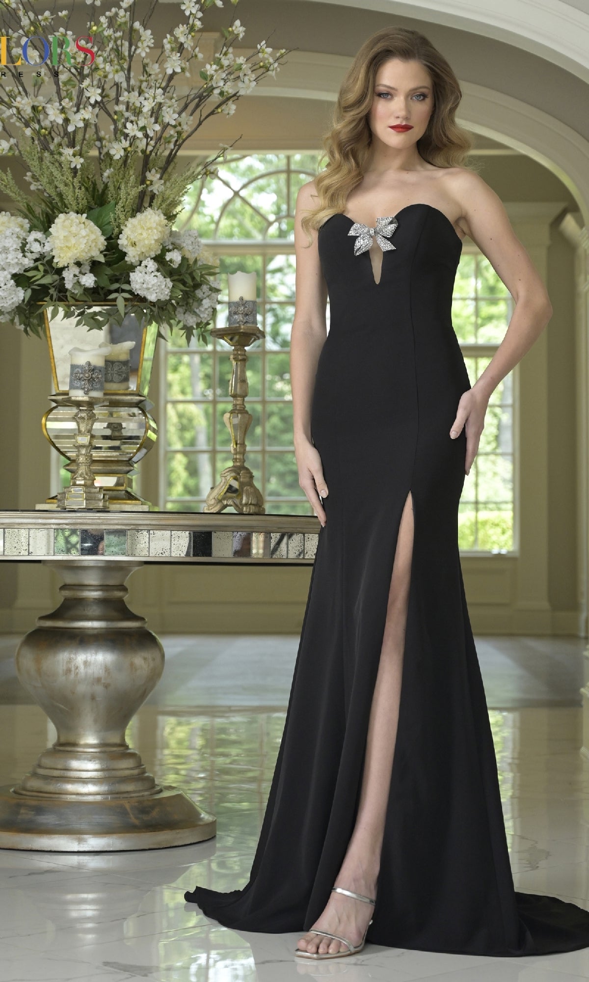 A woman wears the Bow-Back Strapless Prom Gown (Colors Dress 3889), a black strapless long dress with a front slit and bow detail, while standing indoors next to a table decorated with flowers and candles.